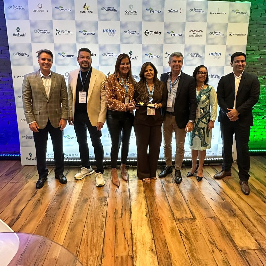 Sete pessoas diante do painel do Kick Off de Distribuidores Sysmex, segurando o troféu da Categoria Ouro